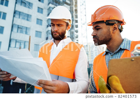 Two young male engineers in uniform and hardhats working at construction site Two young male engineers in uniform and hardhats working at construction site 82401573