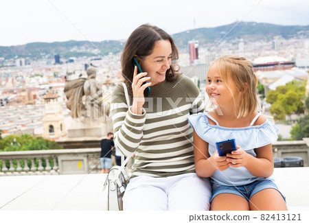 Happy mom with daughter use mobile phones while sitting on parapet 82413121