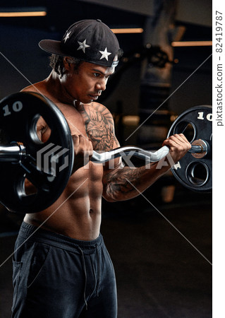 Strong and muscular afro-american man trains on modern equipment in gym. Portrait of muscular pumped up fitness trainer Strong and muscular afro-american man trains on modern equipment in gym. Portrait of muscular pumped up fitness trainer 82419787