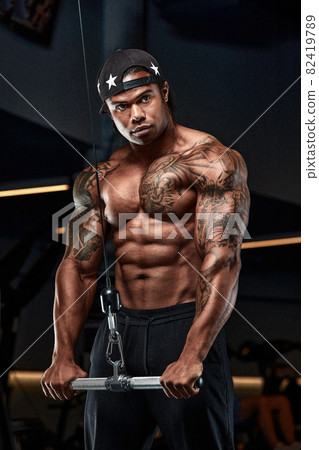 Strong and muscular afro-american man trains on modern equipment in gym. Portrait of muscular pumped up fitness trainer Strong and muscular afro-american man trains on modern equipment in gym. Portrait of muscular pumped up fitness trainer 82419789