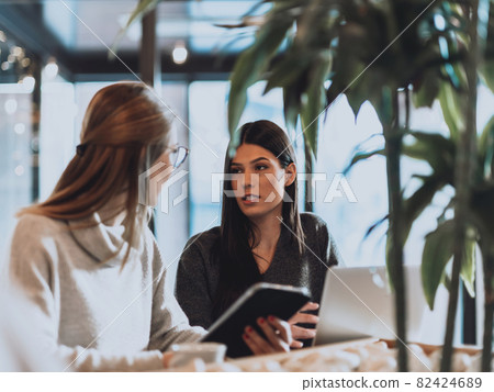 two young business women sitting at table in cafe. Girl shows colleague information on laptop screen. Girl using smartphone, blogging. Teamwork, business meeting.. two young business women sitting at table in cafe. Girl shows colleague information on laptop screen. Girl using smartphone, blogging. Teamwork, business meeting.. 82424689