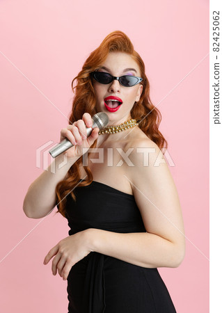 Cropped vertical portrait of red-haired attractive woman in stylish sunglasses singing isolated over pink background Cropped vertical portrait of red-haired attractive woman in stylish sunglasses singing isolated over pink background 82425062
