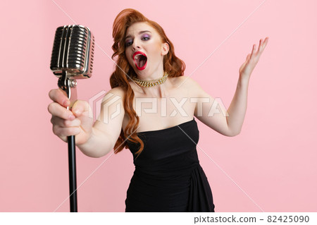 Cropped studio portrait of expressive beautiful woman singing in microphone isolated over pink background. Retro music performer Cropped studio portrait of expressive beautiful woman singing in microphone isolated over pink background. Retro music performer 82425090