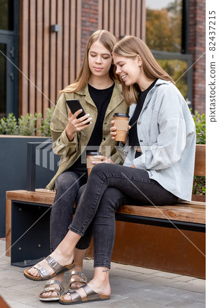 Two blond girls in jeans and jackets sitting on bench and video calling Two blond girls in jeans and jackets sitting on bench and video calling 82437215