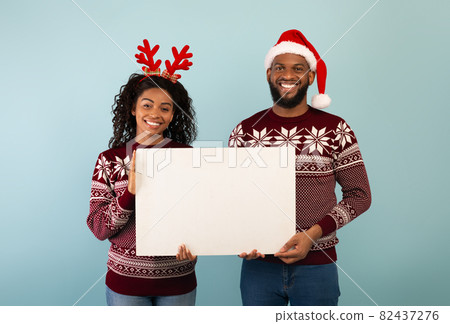 Positive black couple in Christmas outfits holding white poster with empty space, standing over blue background Positive black couple in Christmas outfits holding white poster with empty space, standing over blue background 82437276