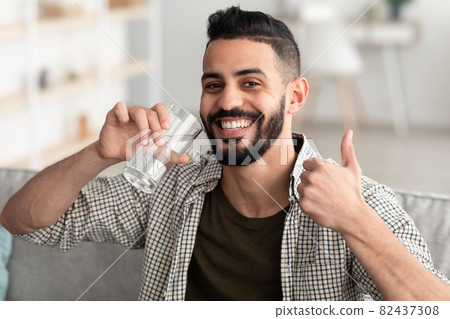 Positive Arab guy drinking fresh clear water from glass, showing thumb up gesture and smiling at camera at home Positive Arab guy drinking fresh clear water from glass, showing thumb up gesture and smiling at camera at home 82437308