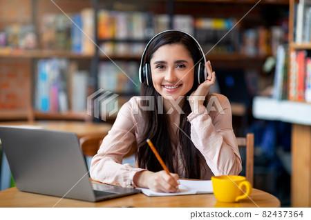 Cheerful latin student in wireless headphones studying online while sitting in cafe, using laptop and taking notes Cheerful latin student in wireless headphones studying online while sitting in cafe, using laptop and taking notes 82437364