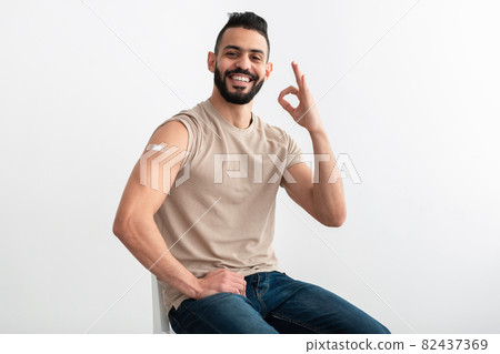 Joyful young Arab guy with vaccinated arm after covid injection showing okay gesture on white studio background 82437369