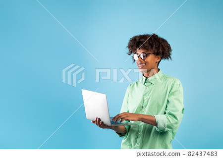 Online education programs. Black teen guy using laptop while standing over blue background, studio shot, free space 82437483