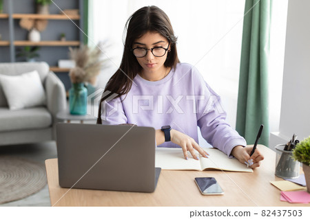 Woman sitting at desk, using pc and writing in notebook 82437503
