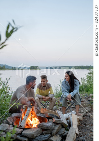Happy family of three sitting by campfire against lake Happy family of three sitting by campfire against lake 82437537