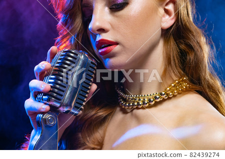 Cropped close-up portrait of young woman, singer with retro hairstyle and gold necklace isolated over gradient blue purple background in neon light 82439274