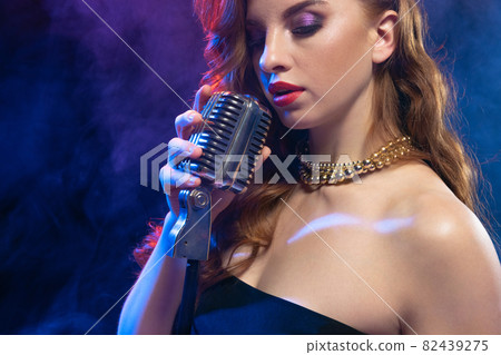 Cropped portrait of young woman, singer with retro hairstyle and gold necklace isolated over gradient blue purple background in neon light 82439275