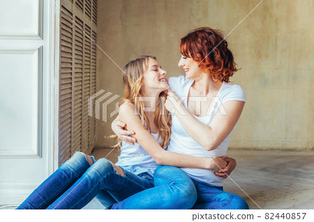 Young mother embracing her child. Woman and teenage girl relaxing in white bedroom near gray wall indoors. Happy family at home. Young mom playing with her daughter. 82440857