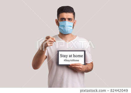 Man in protective mask holding Stay at home inscription and pointing to camera Man in protective mask holding Stay at home inscription and pointing to camera 82442049