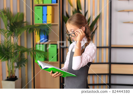 Portrait of a girl in a school uniform with a book in her hands. The child reads a textbook. Preparing a schoolgirl for a school lesson or exam Portrait of a girl in a school uniform with a book in her hands. The child reads a textbook. Preparing a schoolgirl for a school lesson or exam 82444380