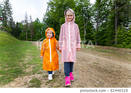 Pair of cute little caucasian sibling kids in bright waterproof raincoats walking under rain in scenic moody dirt forest road outside. Autumn weather outdoor hiking. Children friendship and support 82446328