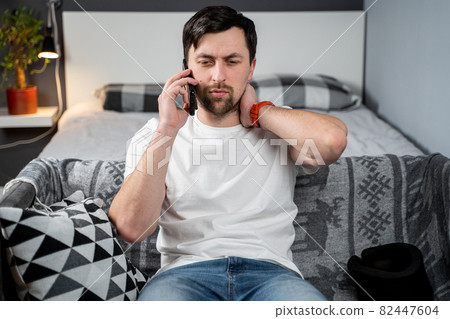 Disabled man with neck brace talking on phone with doctor, experiencing pain while sitting on couch in the living room at home. Injured young man using cervical brace makes calls using mobile phone 82447604