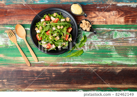 Rocket salad with tomato, crouton, herb roasted chicken in black dish together with wooden utensil and two tiny sauce cups full of mayonnaise and extra crouton on colourful wooden table Rocket salad with tomato, crouton, herb roasted chicken in black dish together with wooden utensil and two tiny sauce cups full of mayonnaise and extra crouton on colourful wooden table 82462149