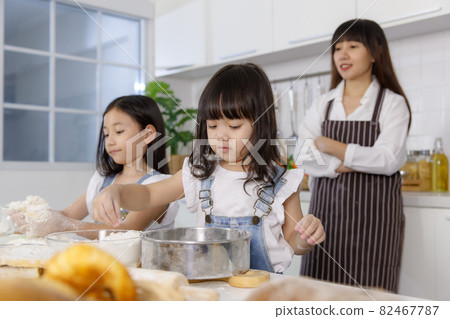 Two little cute Asian girls, 3 years and 7 years old, learning how to make bread and bakery with a curious and happy smiley face. Their mom standing and looking behind them Two little cute Asian girls, 3 years and 7 years old, learning how to make bread and bakery with a curious and happy smiley face. Their mom standing and looking behind them 82467787