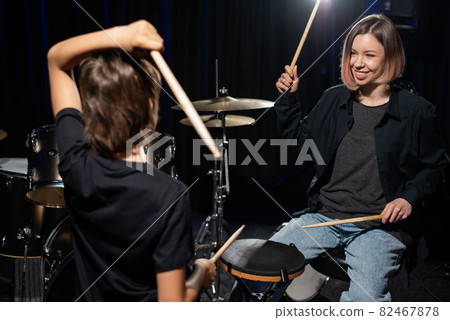 Young caucasian woman teaches a boy to play the drums in the studio on a black background. Music school student Young caucasian woman teaches a boy to play the drums in the studio on a black background. Music school student 82467878