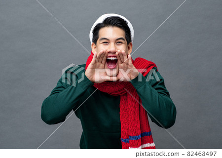 Cheerful young Asian man wearing Christmas attire shouting with hands cupping around mouth in gray  82468497