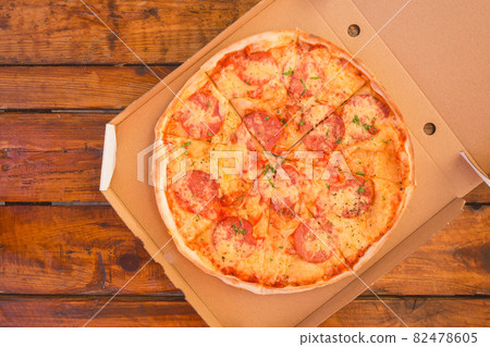 Freshly made pizza with cheese and salami in a cardboard package is a harmful fast food against the background of a wooden table 82478605