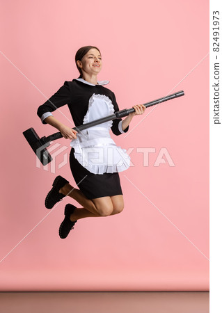 Full-length ortrait of young beautiful chambermaid in a jump holding vaccum cleaner like guitar isolated over pink background. 82491973