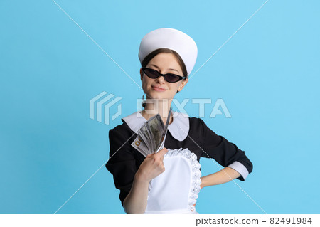 Cropped portrait of young woman in chambermaid uniform and stylish sunglasses with a wad of money isolated over blue background Cropped portrait of young woman in chambermaid uniform and stylish sunglasses with a wad of money isolated over blue background 82491984
