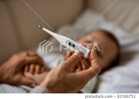 Worried young mother sitting on sofa beside her sick son with high fever. Mom measures temperature 82500751