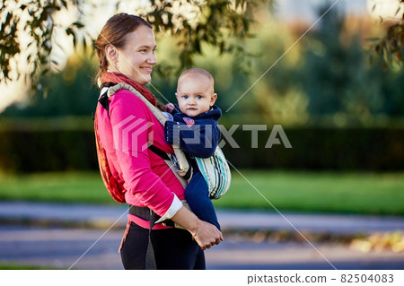 Happy european woman with little boy in sling walks in park. Happy european woman with little boy in sling walks in park. 82504083