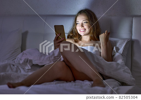 Bare happy woman in bed lies with phone in hand. Bare happy woman in bed lies with phone in hand. 82505304