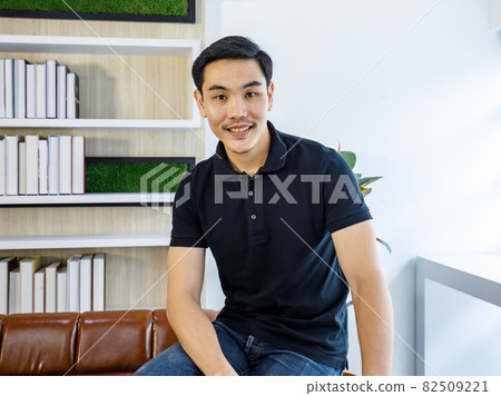 Smart young Asian man in black casual shirt confidently sit at ease on sofa in reading room near book shelf smiling as happy for healthy life in holiday relaxing Smart young Asian man in black casual shirt confidently sit at ease on sofa in reading room near book shelf smiling as happy for healthy life in holiday relaxing 82509221