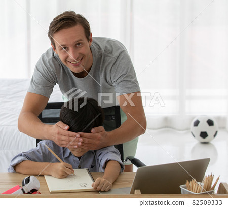 Father with smile and fun face using hand to close eyes of mixed race son who sitting on desk Father with smile and fun face using hand to close eyes of mixed race son who sitting on desk 82509333