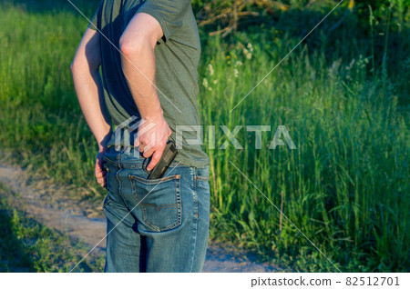 Man in green cloth holds firearm behind his back. Revolver in men's hand. Criminal concept. Person ready to attack with hand gun. Man in green cloth holds firearm behind his back. Revolver in men's hand. Criminal concept. Person ready to attack with hand gun. 82512701
