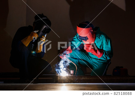 engineer working at night time in a factory's workshop. Two mechanics wearing mechnic jumpsuits, glove and put on a welding helmet working on a metalwork with a welding machine engineer working at night time in a factory's workshop. Two mechanics wearing mechnic jumpsuits, glove and put on a welding helmet working on a metalwork with a welding machine 82518088
