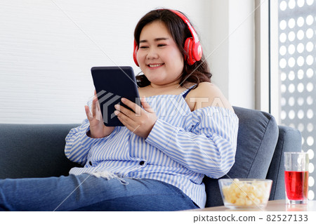 Asian young happy pretty friendly fat plus size woman sit smiling relaxing on gray fabric couch listening to music from big red headphones use playlist from black tablet on hand in living room 82527133