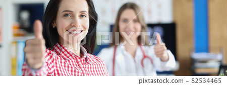 Satisfied healthy smiling young adult female patient showing thumbs up gesture together with doctor Satisfied healthy smiling young adult female patient showing thumbs up gesture together with doctor 82534465