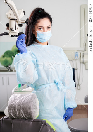 Portrait of a pretty woman dentist, wearing doctor's uniform, face mask, gloves standing in her fully equipped dental office. Modern technologies in stomatology. Concept of dentistry and healthcare 82534790
