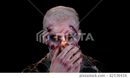 Man with Halloween zombie bloody wounded makeup, trying to scare, spells conjures over a candle 82536416