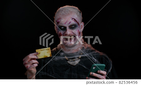 Sinister man Halloween zombie using credit bank card and smartphone while purchases online shopping Sinister man Halloween zombie using credit bank card and smartphone while purchases online shopping 82536456