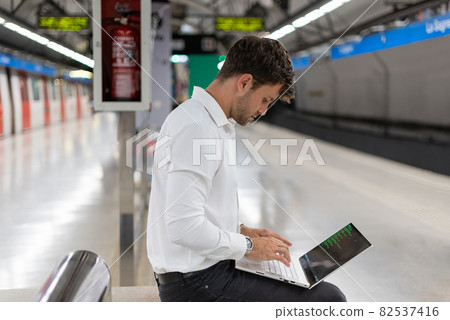 Side view of male in smart casual clothes sitting on bench and typing on netbook keyboard while waiting train on underground station 82537416