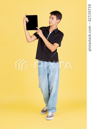 Portrait of young boy, teen or student in casual clothes, black shirt and blue jeans using tablet isolated on yellow studio backgroud. 82539739