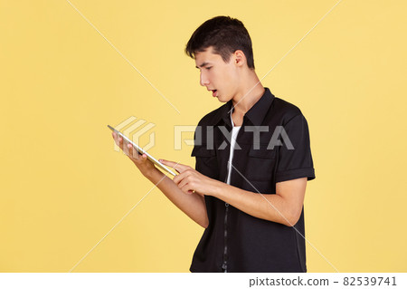 Portrait of young boy, teen or student in casual clothes, black shirt and blue jeans using tablet isolated on yellow studio backgroud. 82539741