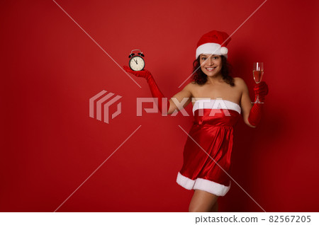 Attractive muscular build young mixed race woman in Santa attire, poses with glass of champagne and an alarm clock, smiles toothy smile, on red background with space for Christmas advertising 82567205