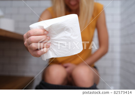 Woman is sitting on t toilet and holding roll of toilet paper 82573414