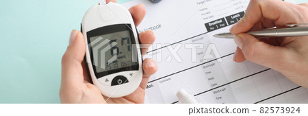 Person holds glucometer with blood sugar readings and makes notes in notebook 82573924