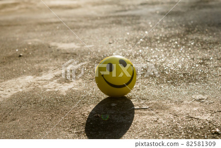 The iconic yellow smiley face with its perfect circle, two oval eyes and a large semi-circular mouth representing a symbol of happiness isolated on sand and shadow background. Happy smile backgrounds. The iconic yellow smiley face with its perfect circle, two oval eyes and a large semi-circular mouth representing a symbol of happiness isolated on sand and shadow background. Happy smile backgrounds. 82581309