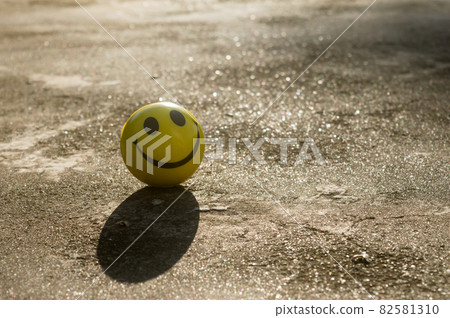The iconic yellow smiley face with its perfect circle, two oval eyes and a large semi-circular mouth representing a symbol of happiness isolated on sand and shadow background. Happy smile backgrounds. The iconic yellow smiley face with its perfect circle, two oval eyes and a large semi-circular mouth representing a symbol of happiness isolated on sand and shadow background. Happy smile backgrounds. 82581310