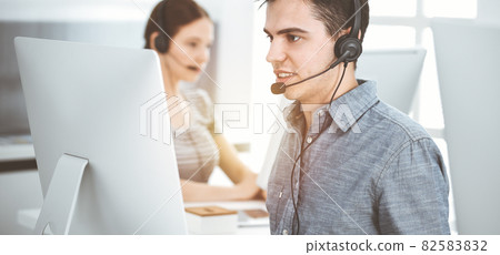 Casual dressed young man using headset and computer while talking with customers online in sunny office. Call center, business concept 82583832